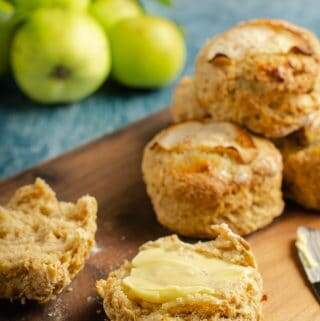 Fresh apple and cinnamon scones piled on a wooden board with freshly picked green apples still with their leaves on just slightly blurred in the background and one scone cut open and topped with salted butter and a butter knife to the side.