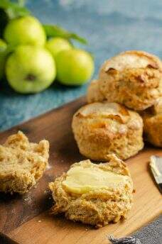 Fresh apple and cinnamon scones piled on a wooden board with freshly picked green apples still with their leaves on just slightly blurred in the background and one scone cut open and topped with salted butter and a butter knife to the side.