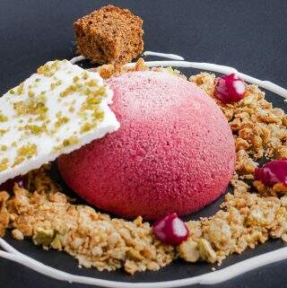 A dome of blackberry parfait served on a matt black plate with oat & pistachio crumb, a swirl of sweetened yogurt, drops of blackberry curd and dark ginger cake topped with a shard of pistachio meringue.