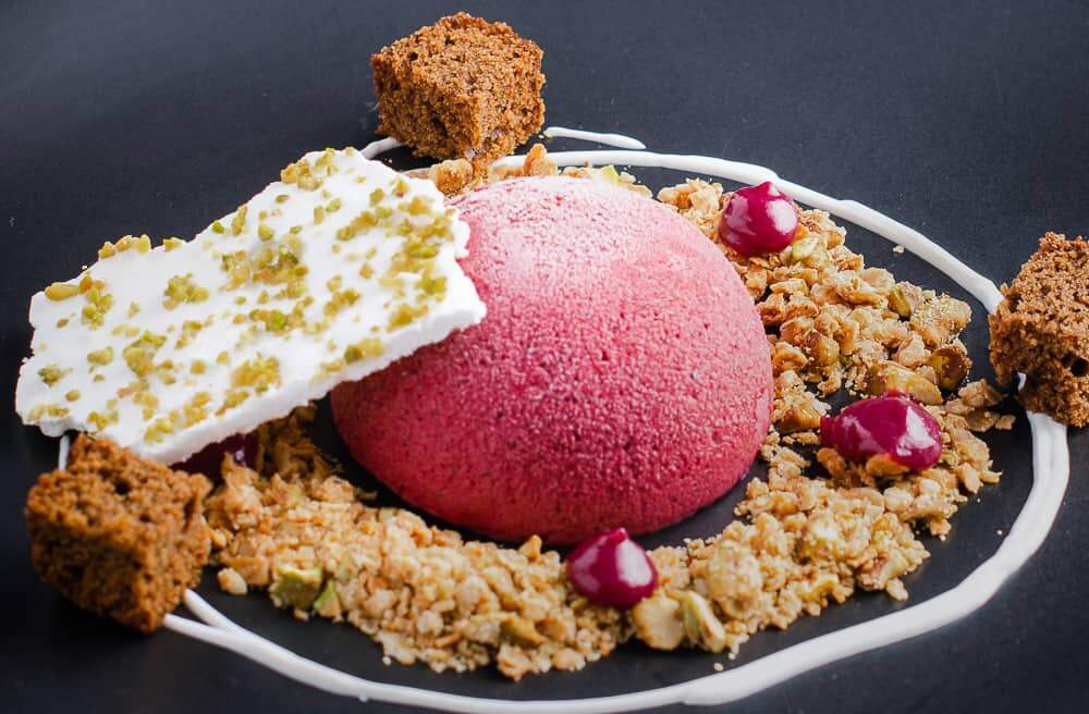 A dome of blackberry parfait served on a matt black plate with oat & pistachio crumb, a swirl of sweetened yogurt, drops of blackberry curd and dark ginger cake topped with a shard of pistachio meringue.