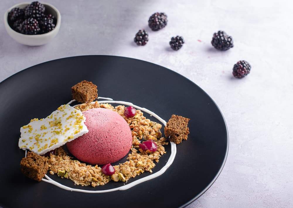 A dome of blackberry parfait served on a matt black plate with oat & pistachio crumb, a swirl of sweetened yogurt, drops of blackberry curd and dark ginger cake topped with a shard of pistachio meringue and a small bowl of fresh blackberries in the back and a few berries loose.