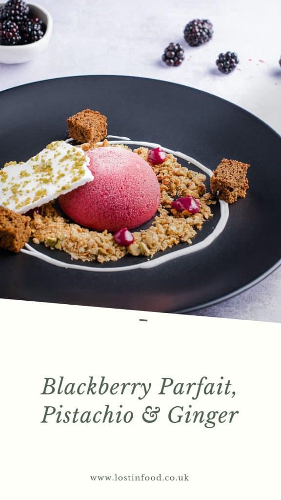 A blackberry parfait dessert served with oat and ginger crumb, a swirl of yogurt, some blackberry curd, fresh ginger cake and topped with pistachio meringue on top of a white border with the dessert name.