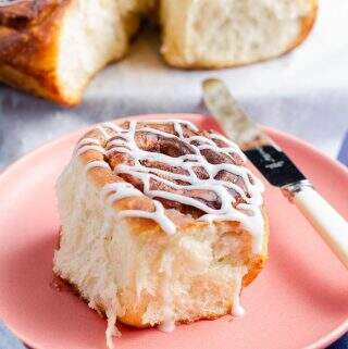 A light an fluffy cinnamon bun sat on a pink plate with a butter knife to the side, drizzled with icing and the remainder of the tray of buns to the back slightly blurred.