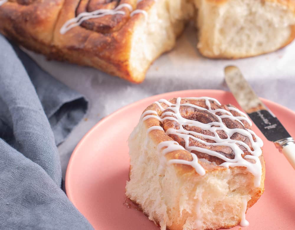 A light an fluffy cinnamon bun sat on a pink plate with a butter knife to the side, drizzled with icing and the remainder of the tray of buns to the back slightly blurred.