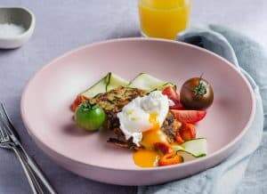 A brunch setting of 2 small gluten free courgette fritters surrounded by fresh tomatoes, slices of courgette and a perfectly poached and runny egg with a glass of orange juice in the back served in a shallow pink bowl and a pale blue napkin to the side and a small pinch pot of salt to the back