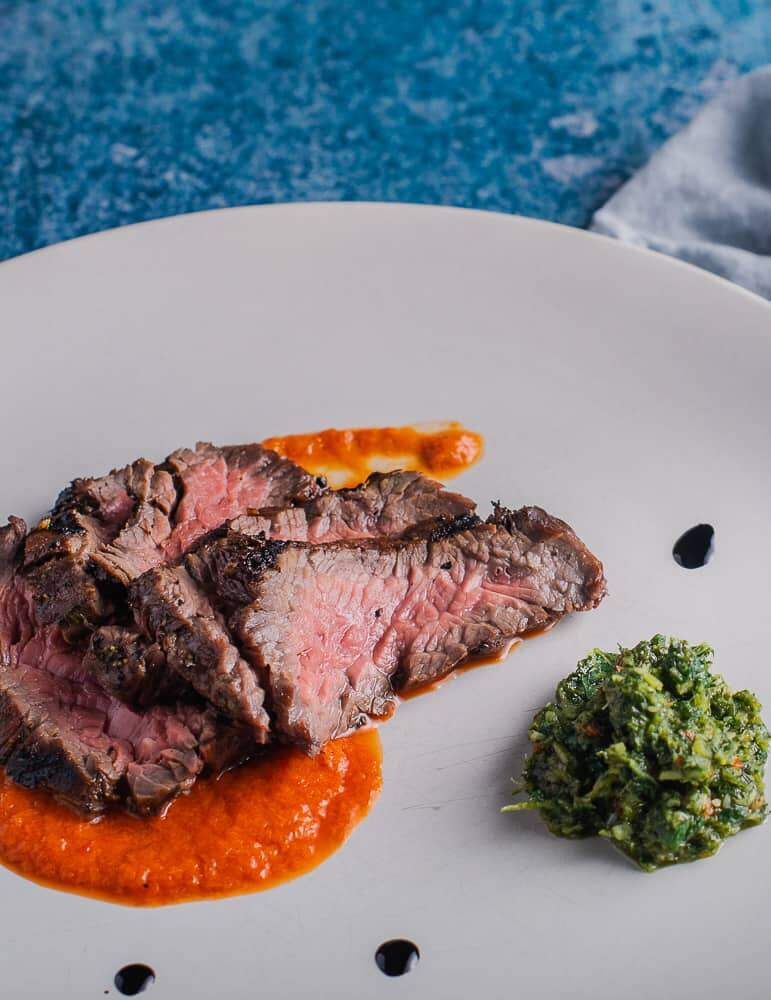 Slices of marinated bavette steak cooked medium rare on a bed of red pepper puree and a side of salsa verde with a pale blue napkin and steak knife in the image.
