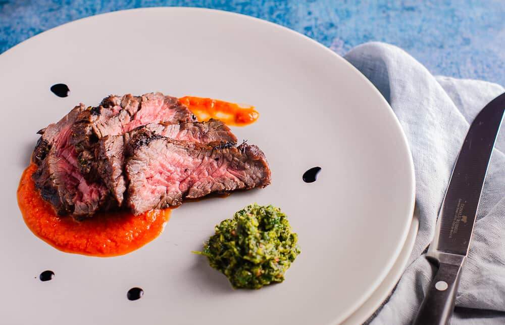 Slices of marinated bavette steak cooked medium rare on a bed of red pepper puree and a side of salsa verde with a pale blue napkin and Donald Russell steak knife in the image.