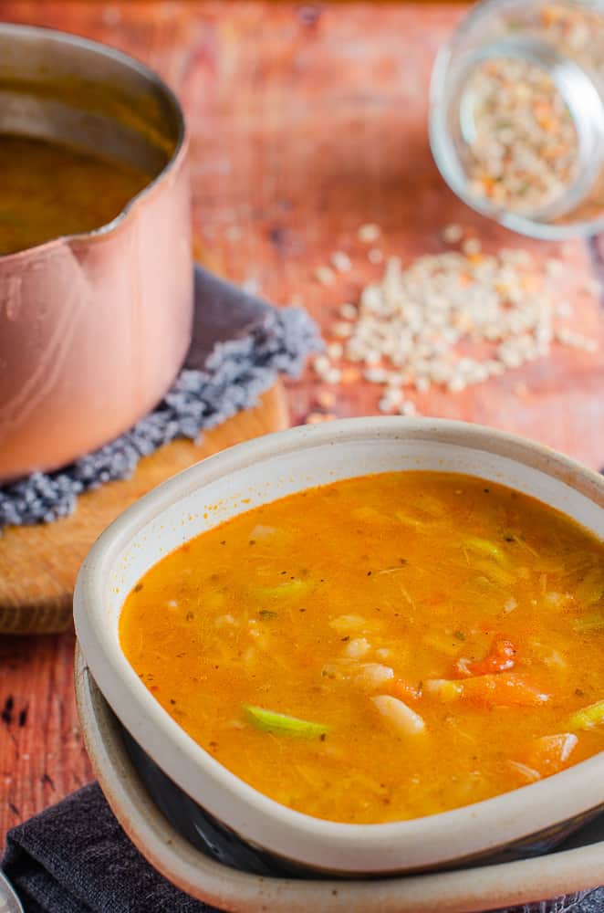 A pottery bowl at the front filled with a leek & barley broth sitting on a navy blue napkin, a copper pan to the side filled with soup on top of a wooden board and a jar of spilled broth mix to the back, all on an old wooden table top.