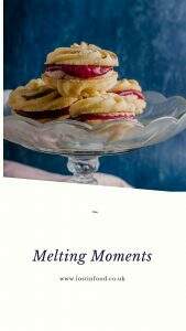 Light buttery biscuits sandwiched together with blackberry curd and buttercream on top of a glass cake stand in front of a blue mottled backdrop with a banner on the bottom that reads melting moments and lost in food, the author.