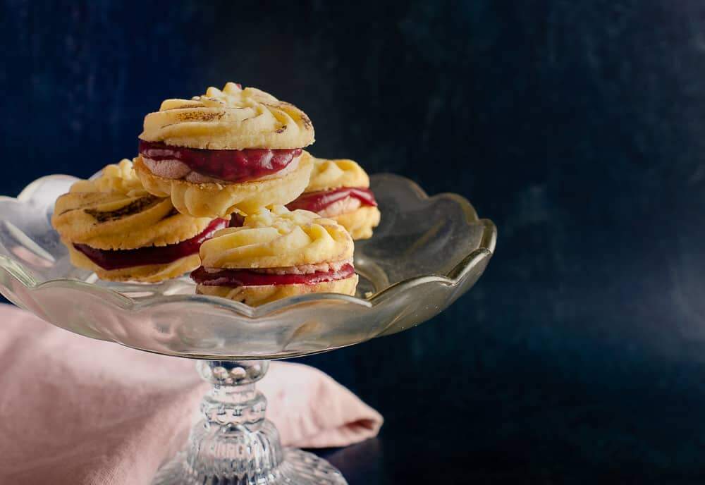 Light buttery biscuits sandwiched together with blackberry curd and buttercream on top of a glass cake stand in front of a dark blue backdrop and a pale pink napkin underneath.