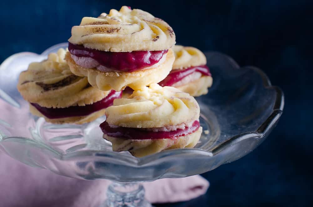 Light buttery melting moment biscuits sandwiched together with blackberry curd and buttercream on top of a glass cake stand in front of a blue mottled backdrop.