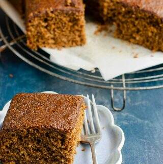 A generous piece of Yorkshire parkin on a small scalloped edge blue ceramic plate with a fork on the side of the plate and the rest of the parkin on a trivet slightly blurred in the background.