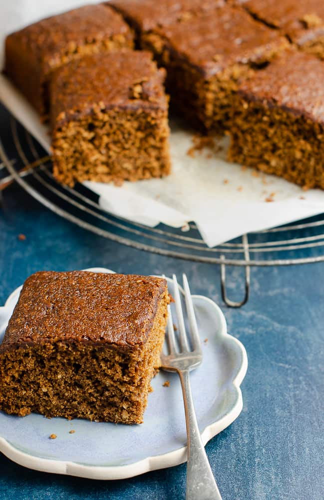 A generous piece of Yorkshire parkin on a small scalloped edge blue ceramic plate with a fork on the side of the plate and the rest of the parkin on a trivet slightly blurred in the background.