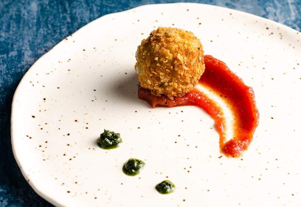 Deep fried potato & cheese ball served on top of a rich tomato sauce with dollops of basil puree on a ceramic plate.