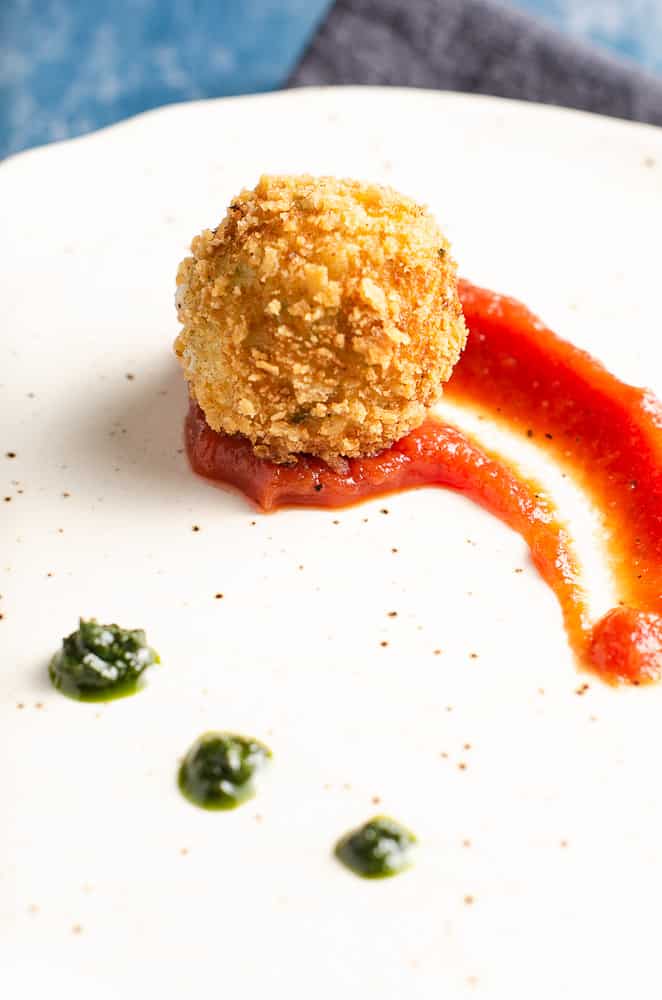 Deep fried potato & cheese ball served on top of a rich tomato sauce with dollops of basil puree on an off white ceramic plate with specs of brown.