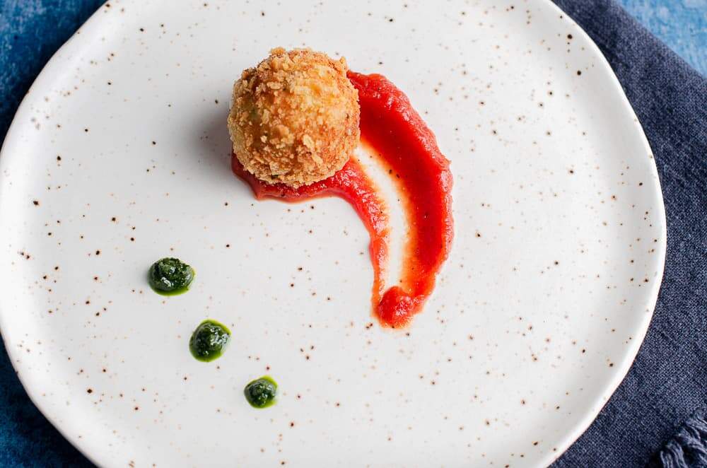 A top down view of a deep fried potato & cheese ball served on top of a rich tomato sauce with dollops of basil puree on a ceramic plate resting on a denim blue linen napkin.