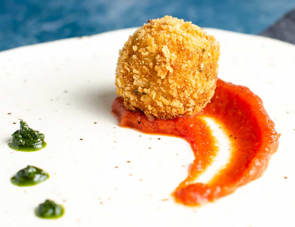 A Closeup view of a deep fried potato & cheese ball served on top of a rich tomato sauce with dollops of basil puree on a ceramic plate with specks of brown and a blue backdrop.