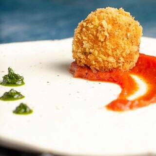 A front close up view of a deep fried potato & cheese ball served on top of a rich tomato sauce with dollops of basil puree on an off white ceramic plate with specs of brown.