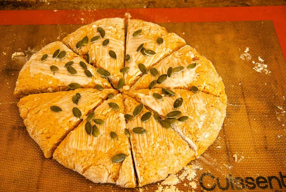One large pumpkin spice scone mix formed into a round and then sliced like a pizza into 8 potions, brushed with leftover buttermilk and topped with pumpkin seeds ready for the oven to be backed.