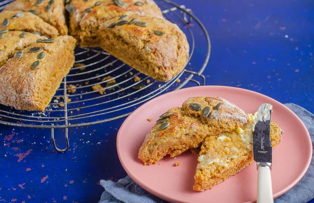 Pumpkin spice scones, one in the foreground cut open and spread with butter on a pink plate and the remaining batch in the back on a old round wire trivet and sitting on a dark blue backdrop with orange flecks.