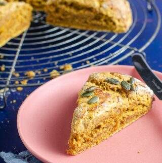 A bright pink plate with a wedge of pumpkin spice scone on top of blue napkins and a dark blue backdrop with a cooling rack filled with the remaining batch of scones.