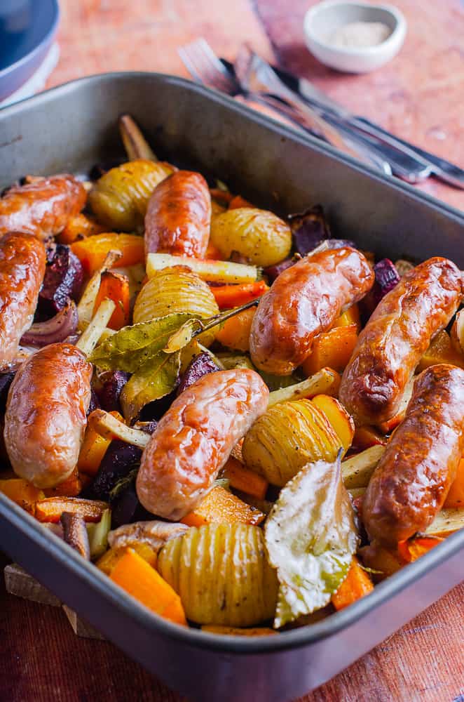 A sticky sausage traybake consisting of potatoes, red onions, parsnips, beetroot, carrot and squash topped with fresh bay and roasted serving bowls and a salt dish behind.