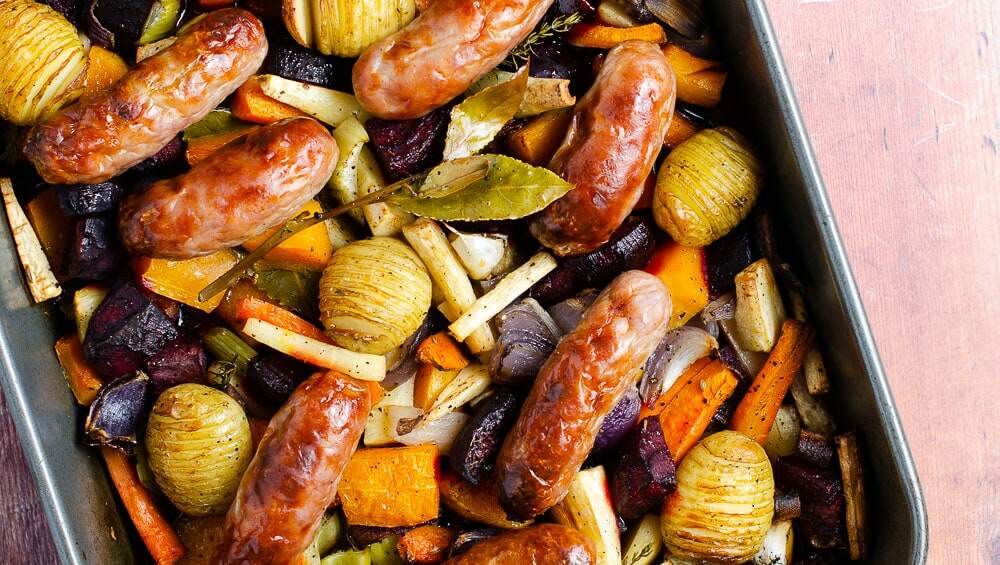 A sticky sausage traybake from above consisting of potatoes, red onions, parsnips, beetroot, carrot and squash topped with fresh bay and roasted serving bowls and a salt dish behind.