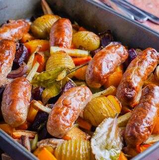 A large pan filled with roasted carrots, squash, parsnips, red onions and sticky coated sausages on an old wooden backdrop ready to eat.