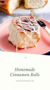 A light an fluffy cinnamon bun sat on a pink plate with a butter knife to the side, drizzled with icing and the remainder of the tray of buns to the back slightly blurred and a white border to the bottom which reads homemade cinnamon rolls and Lost in Food, the Author