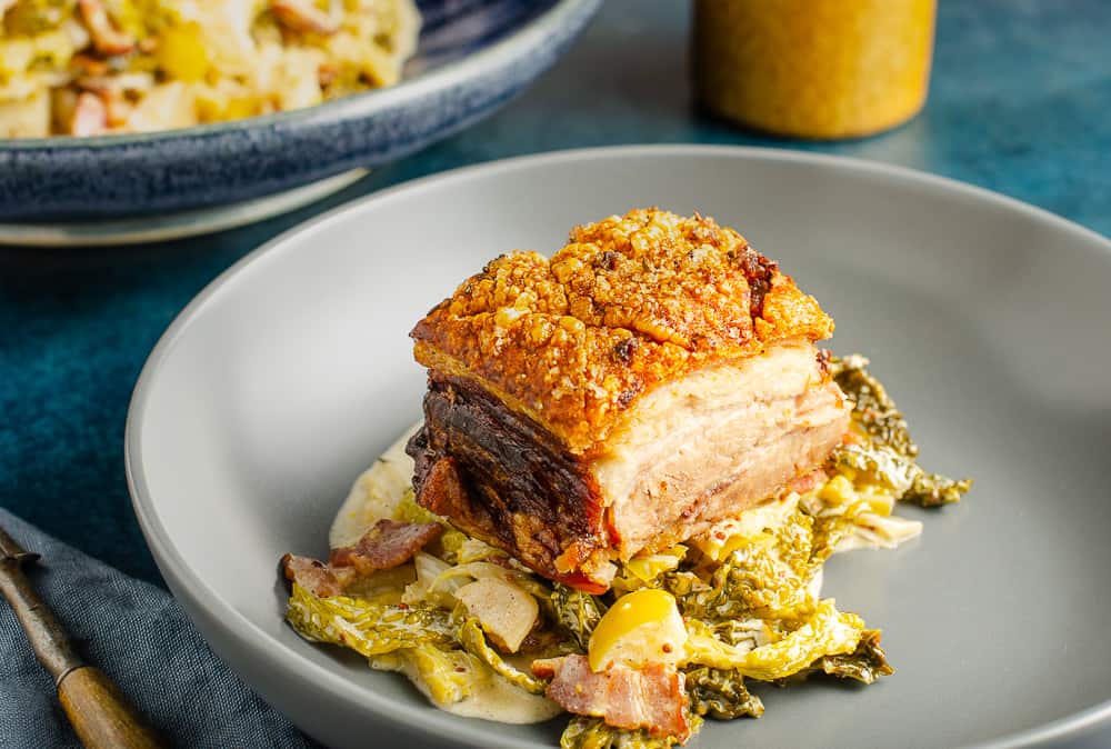 A side dish of cabbage cooked with bacon, apple and cider served under a juicy piece of pork belly and a big serving bowl of the cabbage to the back.