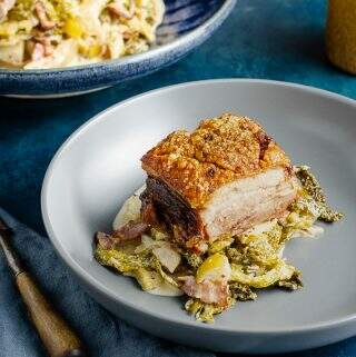 A side dish of cabbage cooked with bacon, apple and cider served under a juicy piece of pork belly and a big serving bowl of the cabbage to the back.