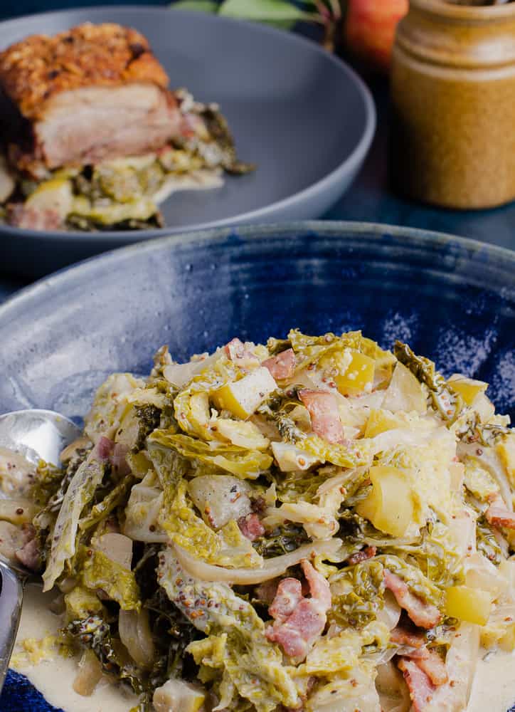 A large serving bowl of creamed cabbage cooked with apples, cider and bacon served alongside pork belly with crispy crackling.