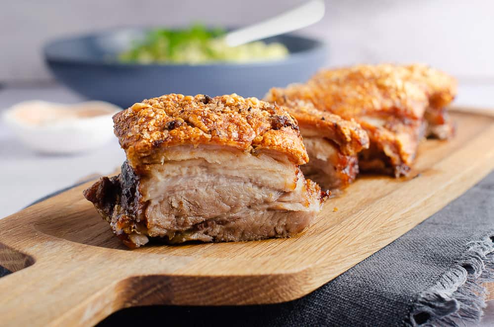 A close up view showing the layers in slices of pork belly, the extra crisp crackling on top and a bowl of vegetables to the back served on a wooden serving board.