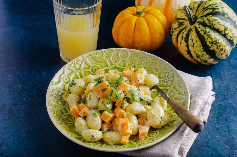 A plate of creamy gnocchi with pan fried pumpkin pieces and sage served an an ornate green plate, with a glass of orange juice in the back and a mix of 3 orange and green pumpkins to the back with a light beige napkin and wooden handle fork in the dish all on top of a dark green mottled surface.
