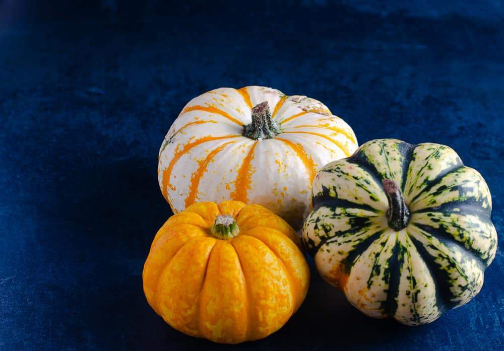 3 pumpkins, a variety of sizes, one bright orange, one white and orange and the other pale green with dark green markings, all sat together on a mottled dark green surface.
