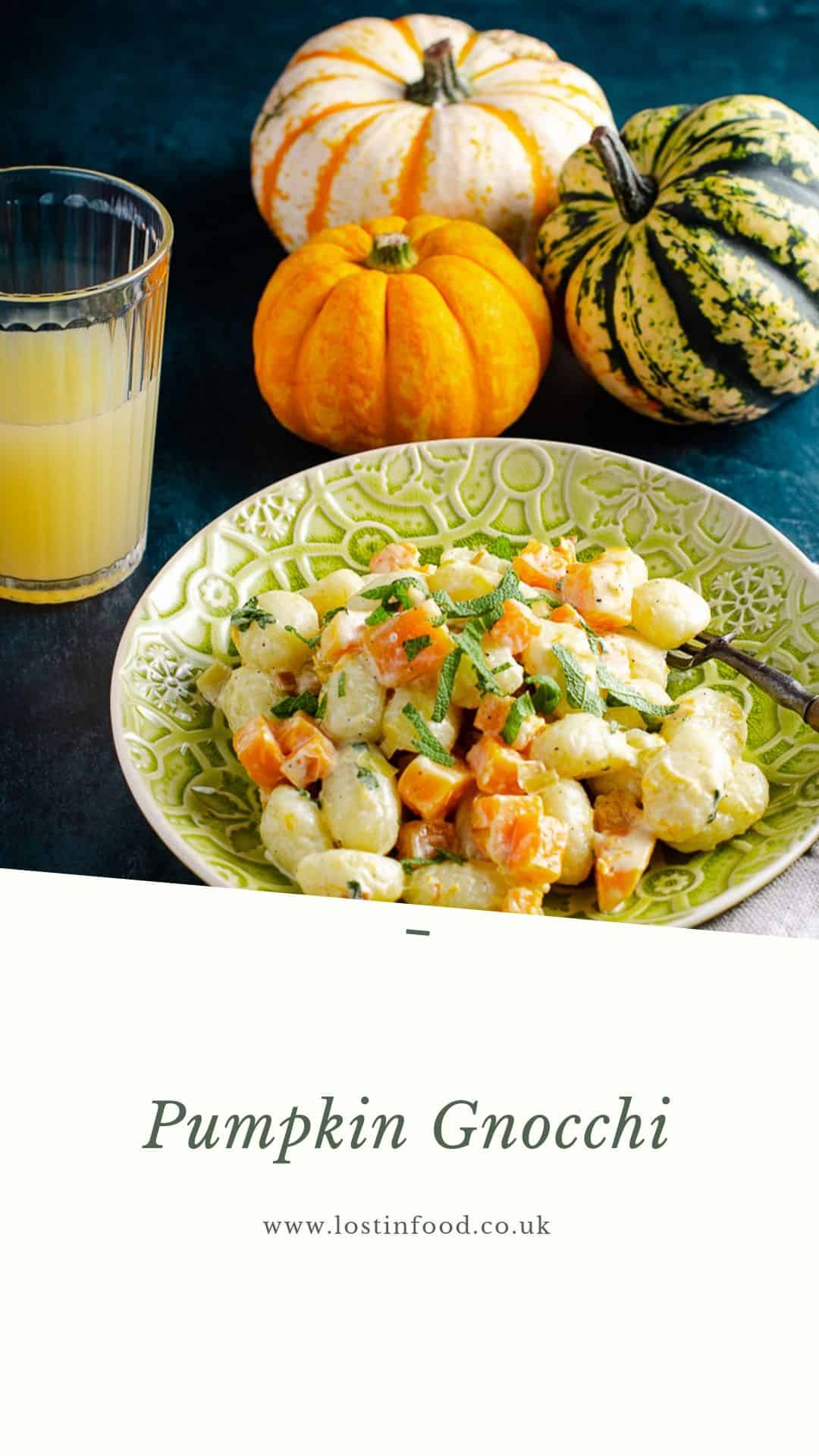 A plate of creamy gnocchi with pan fried pumpkin pieces and sage served an an ornate green plate, with a glass of orange juice in the back and a mix of 3 orange and green pumpkins to the back with a light beige napkin and wooden handle fork in the dish all on top of a dark green mottled surface. A white banner below with the words Pumpkin Gnocchi and Lost in Food as the author.