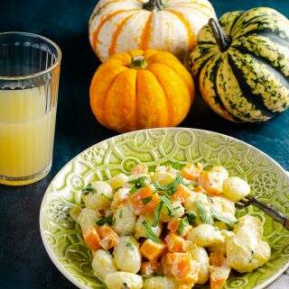 A plate of creamy gnocchi with pan fried pumpkin pieces and sage served an an ornate green plate, with a glass of orange juice in the back and a mix of orange and green pumpkins to the back.