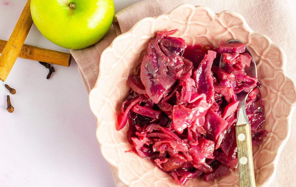 A top down view of a small pink bowl of braised red cabbage, some green apples, cinnamon sticks and cloves on a pink napkin.