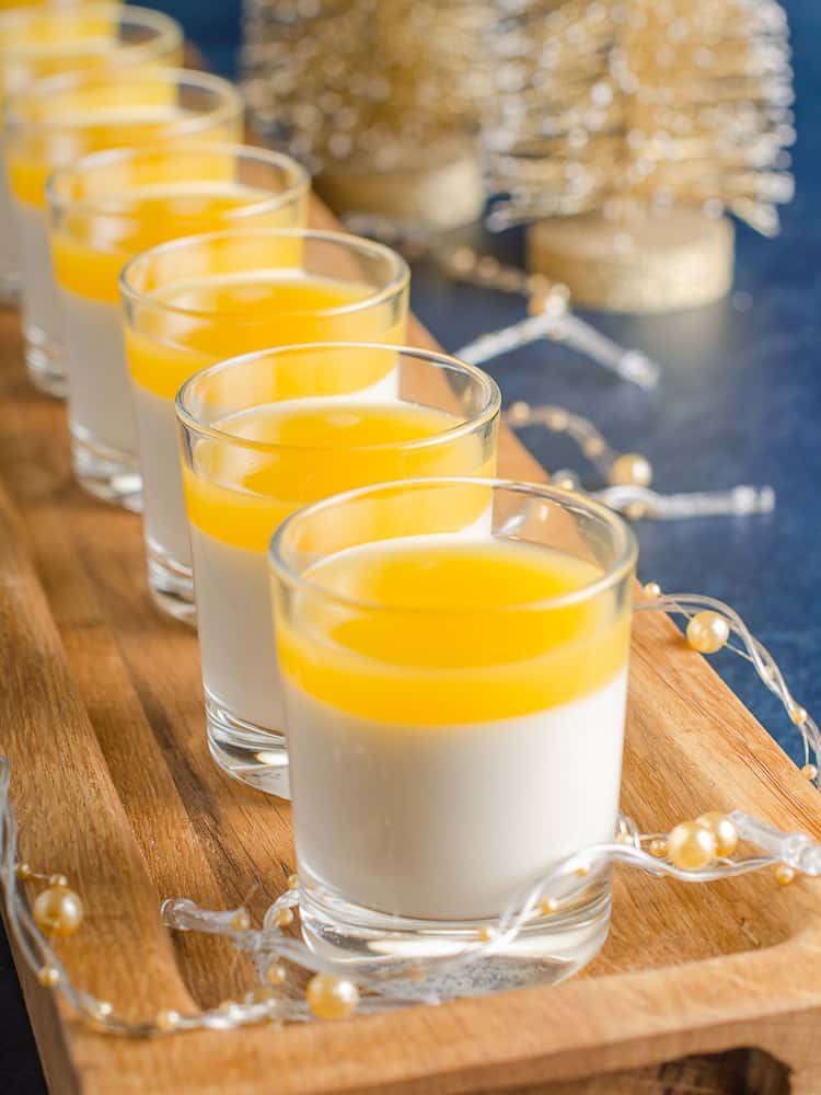 A long wooden serving tray with small individual shot glasses filled with vanilla panna cotta and topped with clementine jelly, with gold Christmas Trees and Golf lights decorating.