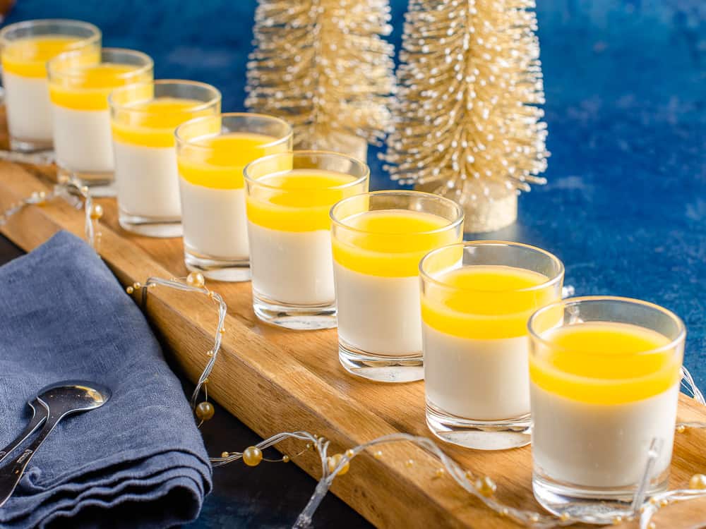 A long wooden serving tray with small individual shot glasses filled with vanilla panna cotta and topped with clementine jeely, with gold Christmas Trees and Golf lights decorating.
