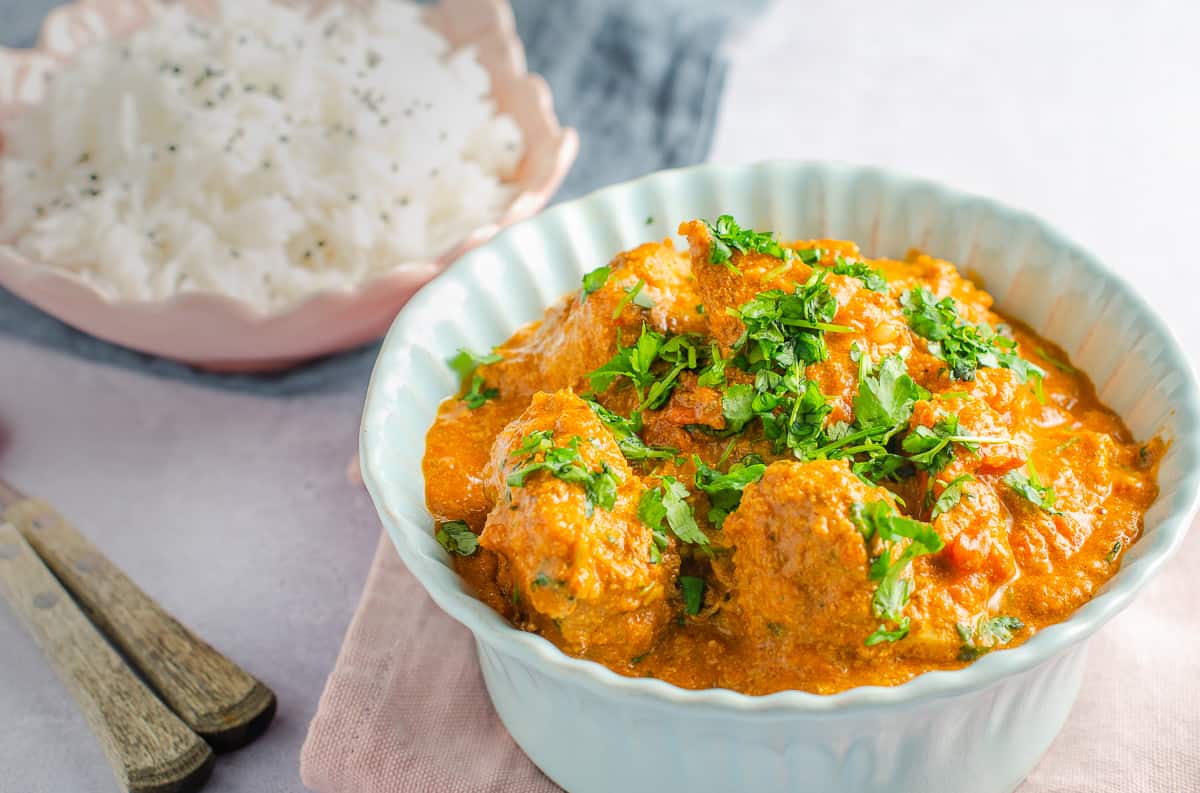 A delicious bowl of butter chicken in a pale blue bowl sat on a pink napkin with a bowl of rice to the back and topped with chopped corriader