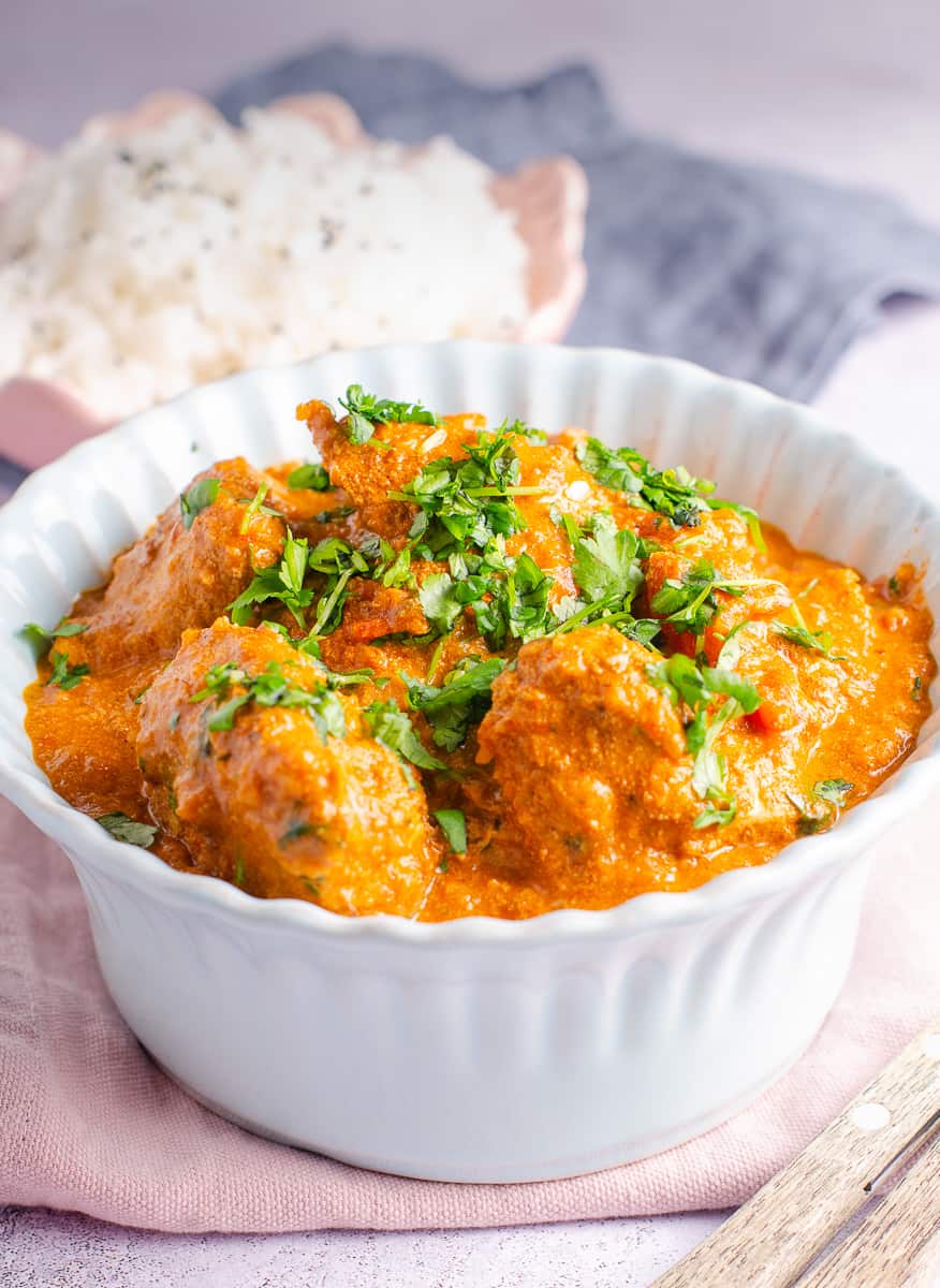 A delicious bowl of butter chicken in a pale blue bowl sat on a pink napkin with a bowl of rice to the back.