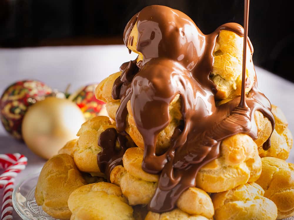 A tower of profiteroles filled with chestnut creme patisserie and warm melted chocolate being poured over the top with some christmas baubles and candy canes in the back