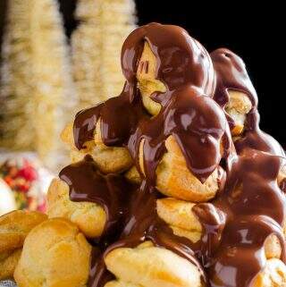 A tower of profiteroles filled with chestnut creme patisserie and warm melted chocolate being poured over the top with some gold christmas trees in the back.