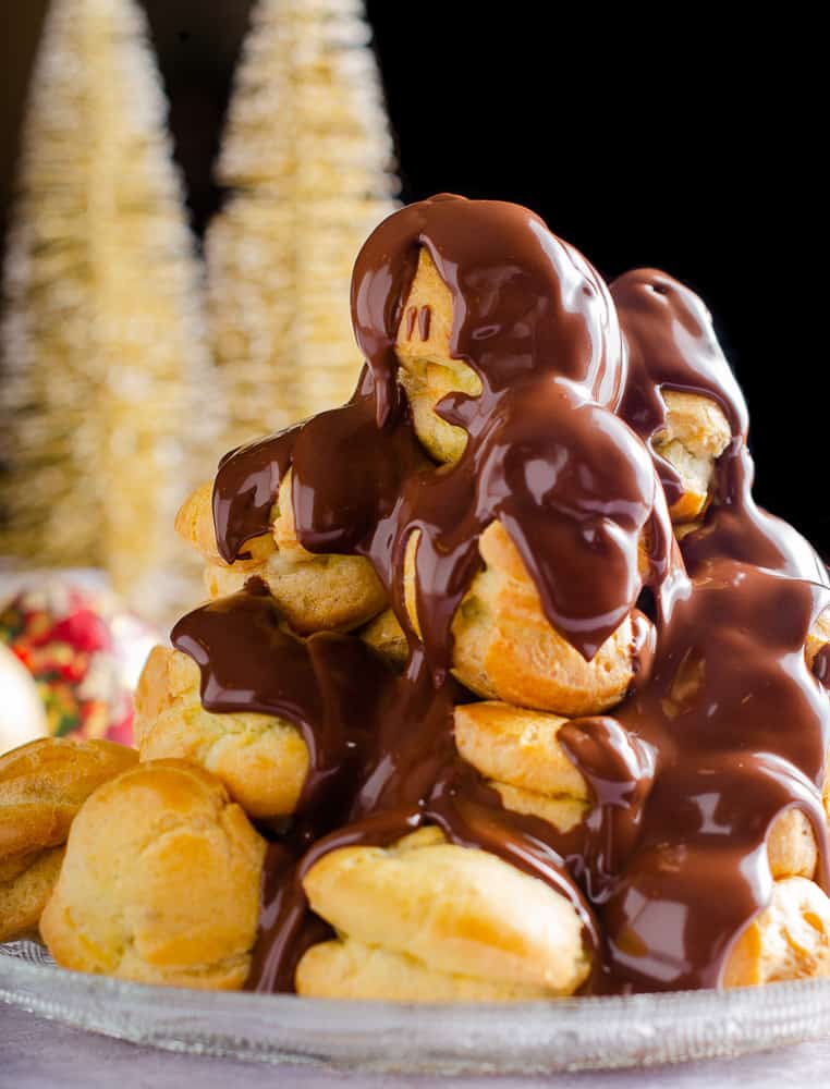 A tower of profiteroles filled with chestnut creme patisserie and warm melted chocolate being poured over the top with some gold christmas trees in the back.