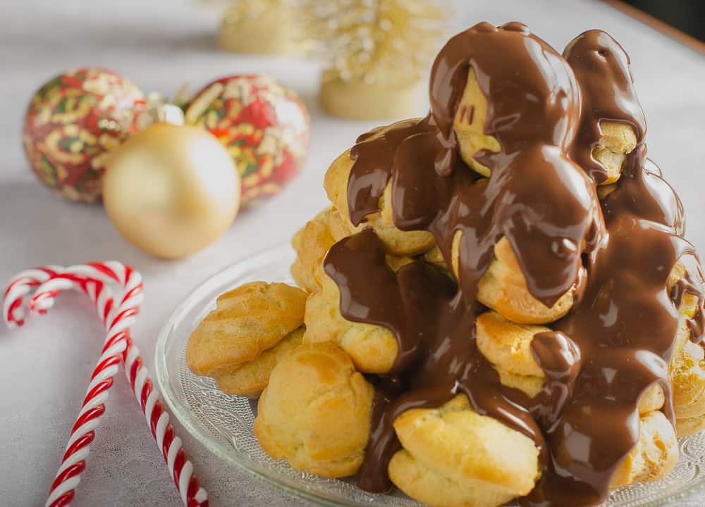 A tower of profiteroles filled with chestnut creme patisserie on a glass plate with some candy canes to the side and christmas baubles to decorate.