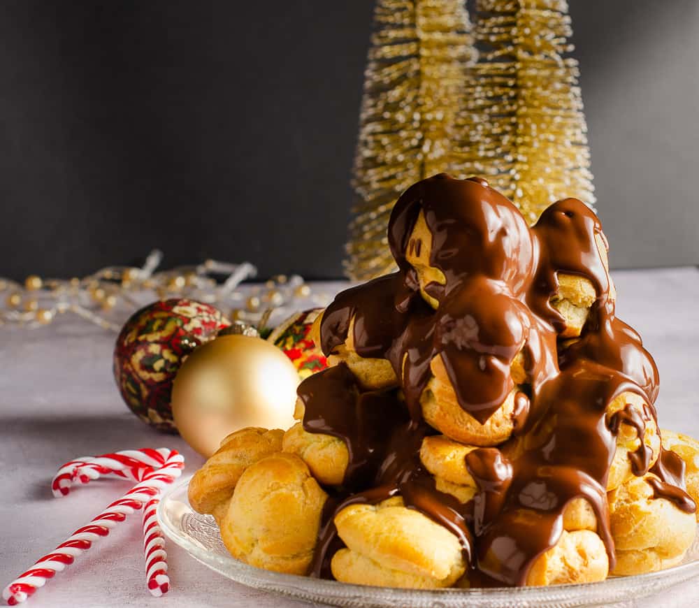 A tower of profiteroles filled with chestnut creme patisserie and warm melted chocolate over the top with some gold christmas trees in the back.