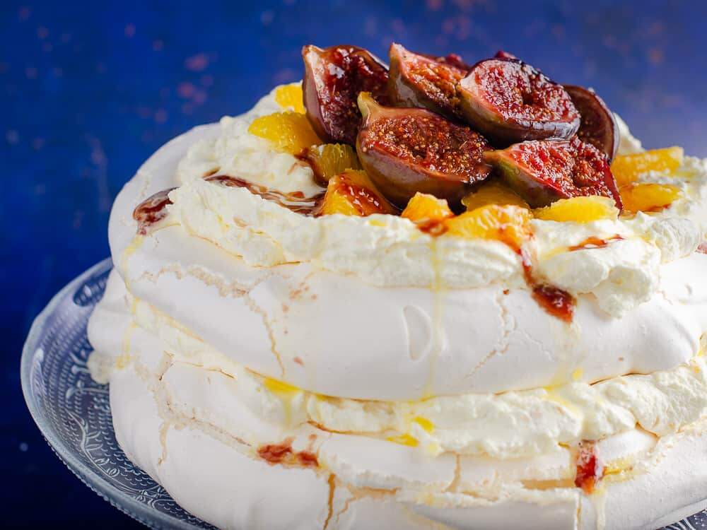 A 2 layer Christmas Pavlova filled with sweetened cream and topped with sticky figs and orange segments on a glass cake stand on a dark blue backdrop.