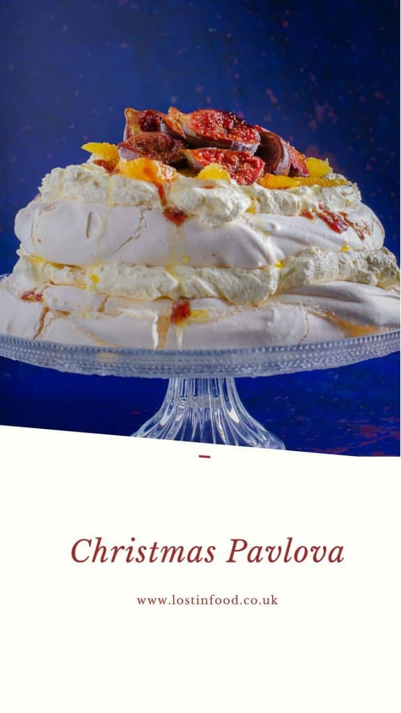 A 2 layer Christmas Pavlova filled with sweetened cream and topped with sticky figs and orange segments on a glass cake stand on a dark blue backdrop and written underneath the name Christmas Pavlova and the authors Lost in Food.