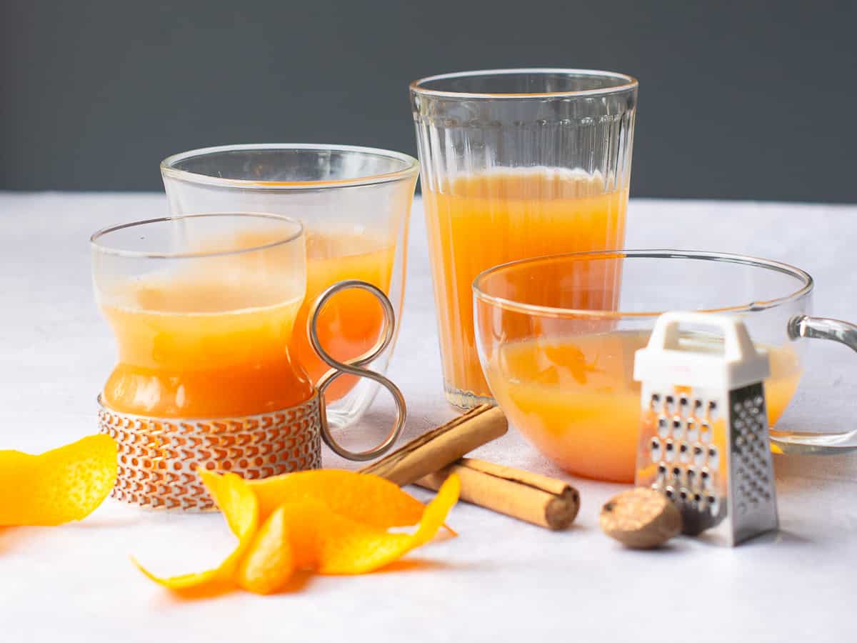 Various glasses filled with mulled apple juice, some orange peels curled, cinnamon sticks and a piece of whole nutmeg with a small nutmeg grater on a pale grey background.